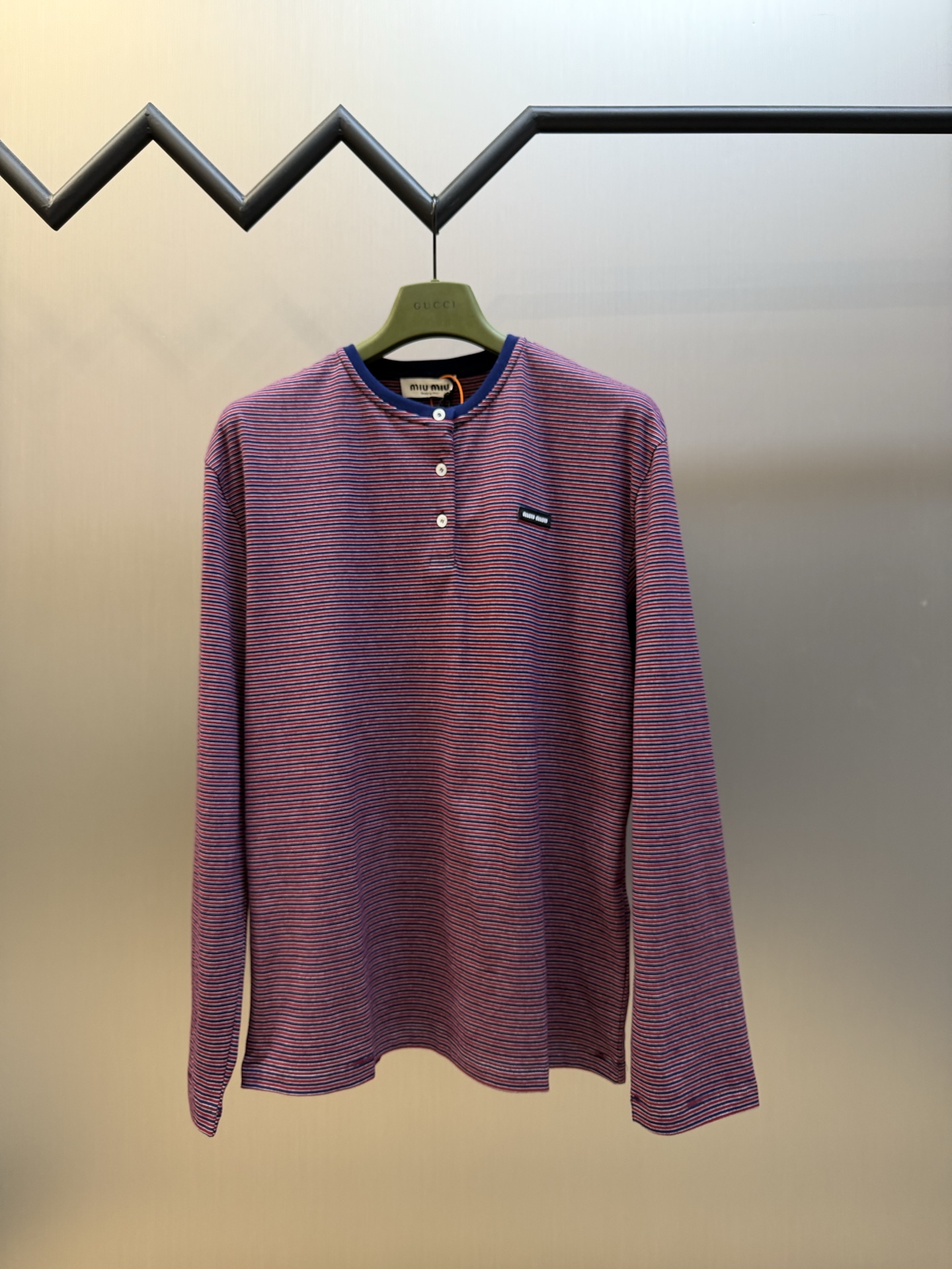 Miu logo striped henley collar long sleeves Customized textured plain weave cotton, intertwined green and burgundy thin strips of warp and weft, layered colors, classic henley half placket configuration, neat opening and closing, contrasting collar edge delicately outlines the collar, brand woven logo sewn on the left chest, loose dropped shoulders silhouette, integrating the Italian retro college heritage into the daily shape, interpreting a relaxed and elegant high-end elegance style