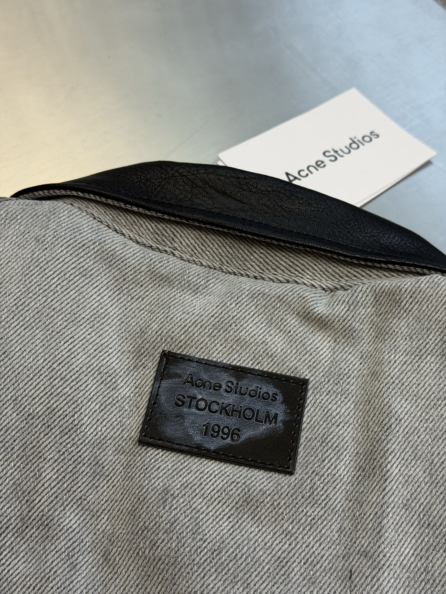 Ac Studios distressed leather cotton denim jacket The outer layer is custom-made weft-ordered organic denim, and the inner layer is custom-made domain-ordered plaid cotton. The entire body is covered in a matte gray tone. The original faded texture is naturally generated. The black leather collar is neatly cut and spliced. The roughness of the denim blends with the smoothness of the leather, creating a collision of rich layers of materials. The arms are embossed with dark patterns, and the patterns are dimly visible. The classic leather label on the back of the neck is properly seated, and the brand's collection imprint is hidden on the back. - Image 7