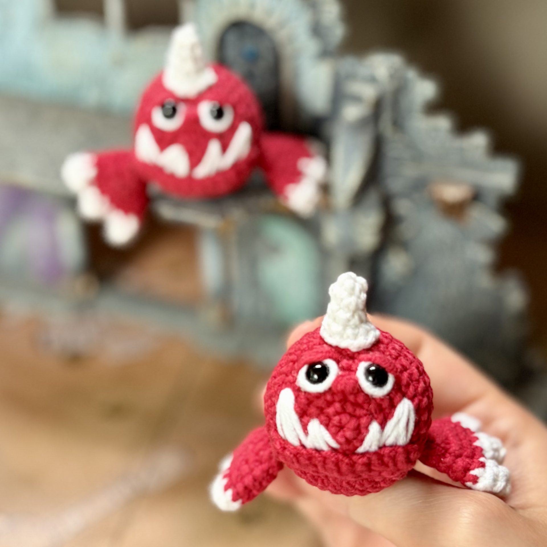 Pocket Squig Crochet Pattern. Warhammer Fantasy Amigurumi PDF 5 Crochet pattern featuring a tiny red Warhammer Squig held in a hand, showing the finished toy's size, with another Squig resting on fantasy ruins in the background