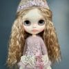 Charming Blythe doll with blond hair and soft pink lips, embodying shy innocence. Dressed in a light pink dress