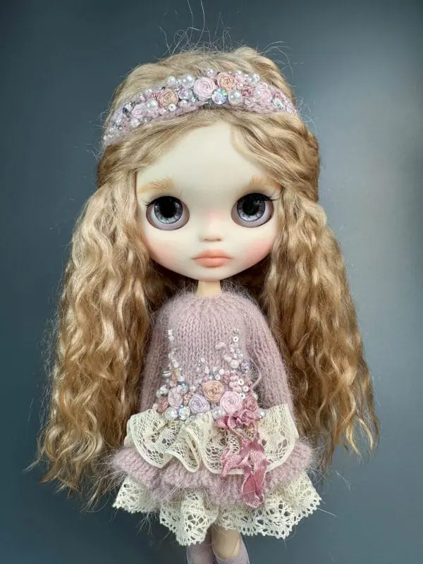 Charming Blythe doll with blond hair and soft pink lips, embodying shy innocence. Dressed in a light pink dress
