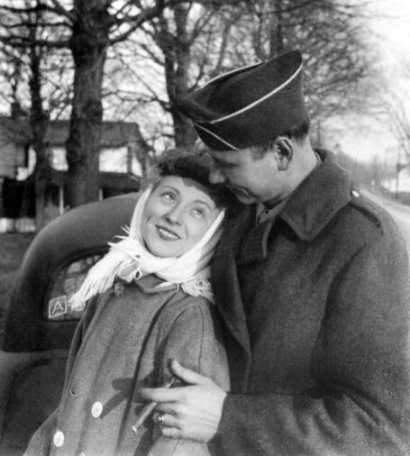 A black and white image of a man and a woman. The man had just returned from wwii. The man is much taller than the woman. He is holding her from behind while she is leaning back and looking up at him and lovingly into his eyes. The man is wearing his military hat and overcoat. The woman is wearing a scarf and a coat. It looks like wintertime. The man is holding a cigar. The people were my wife's parents.There is part of an automobile and trees visible behind them