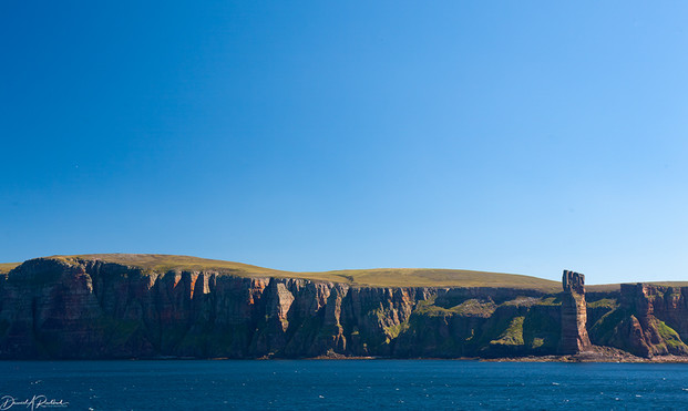 Island coastline seen from sea, with one taller seastack separated from the main landmass. Blue sea and bluer sky
