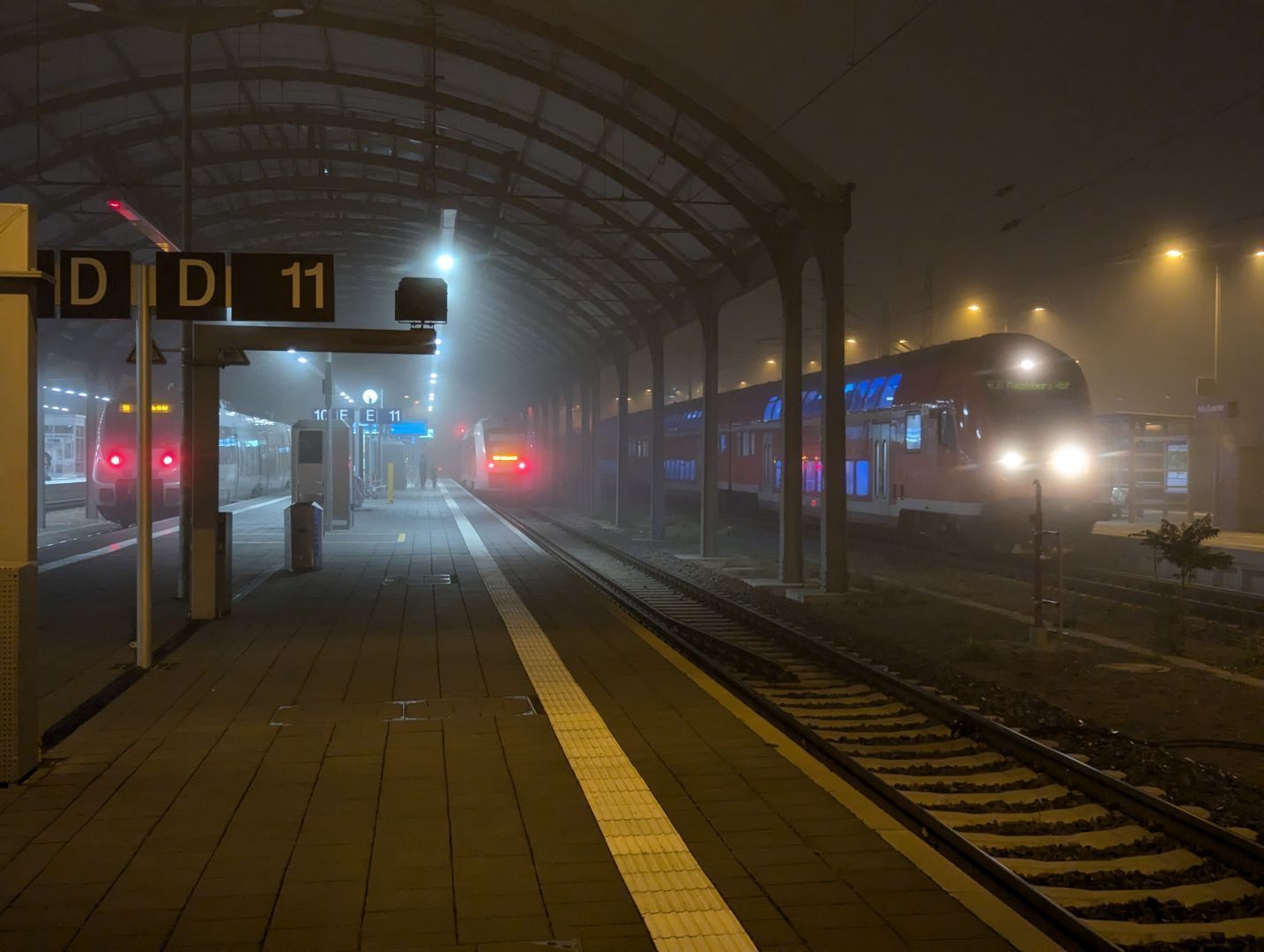 A photo of Halle Hbf from platform 11, just outside the ceiling. A double-decker train is pulling in, and 2 trains are already standing at platforms 11 and 10. One can barely make things out at this distance, except for the close-by double-decker train and the rear end lights of the other 2 trains.