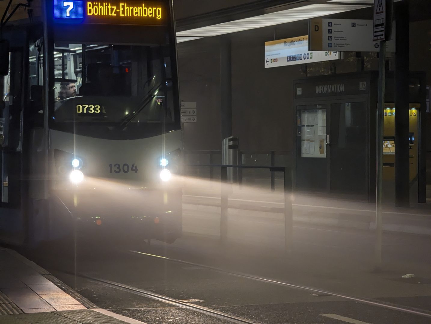 A photo of a HeiterBlick Leoliner (NGT6) tram in Leipzig, specifically Tz 1304, bound to Böhlitz-Ehrenberg as line 7. It's quite dark outside, with some signs visible saying one's at the main station tram stop, and the front lights of the tram cause the fog to cause some nice light diffusion.