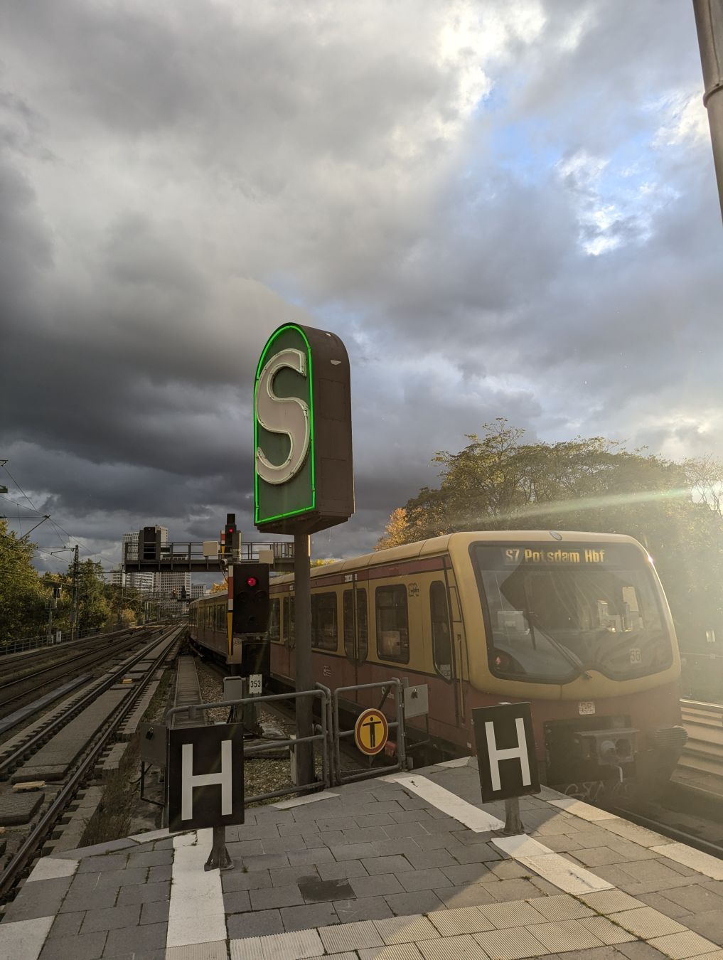 A photo of a neon sign for the S-Bahn in Berlin, with a class 481/482 S-Bahn bound to Potsdam Hbf driving along next to it. The sky is a bit cloudy but the sun was shining in just the right angle to 