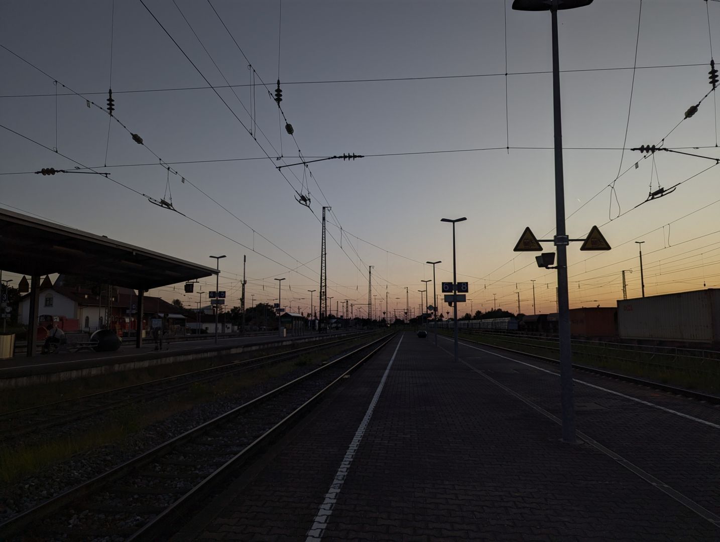 A photo taken from the station platforms 6 and 8 at Plattling. You can see lots of overhead wires, some signs, the platform for the tracks 4 and 5 and a fairly bright sky. As usual, the photos are fairly dark while the sky is still fairly bright.