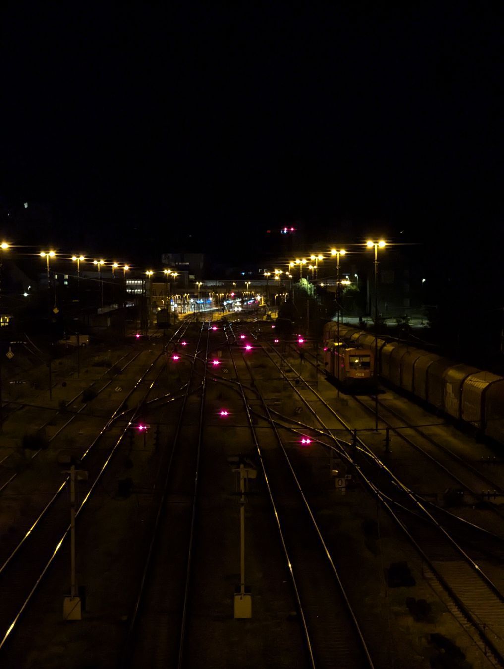 A photo of the tracks leading towards Passau Hbf at night. You can see two rows of lights standing between some off-site tracks, and an ÖBB Taurus loco next to freight carriages. Further to the back, you can partially see the train station.