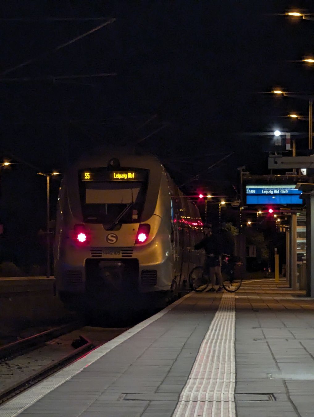 An S5 towards Leipzig Hbf is waiting for passengers to board the train in Halle (Saale) Hbf. People with bicycles are boarding it. The departure board is saying that it leaves at 23:59.
The image is fairly dark, with only parts of the platform being lit (partially because it was in the hall still, partially because of those kinds of lights you'd typically see in street lights lighting up the platform).